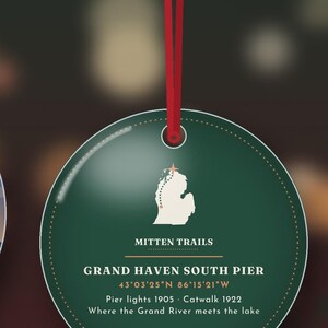 May include: A round, dark green Christmas ornament hanging from a red ribbon. It features a white map of Michigan with a star, the text MITTEN TRAILS, GRAND HAVEN SOUTH PIER, coordinates, and the dates Pier lights 1905 and Catwalk 1922.