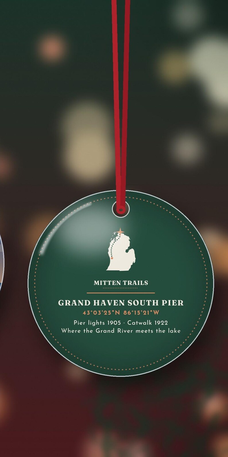 May include: A round, dark green Christmas ornament hanging from a red ribbon. It features a white map of Michigan with a star, the text MITTEN TRAILS, GRAND HAVEN SOUTH PIER, coordinates, and the dates Pier lights 1905 and Catwalk 1922.