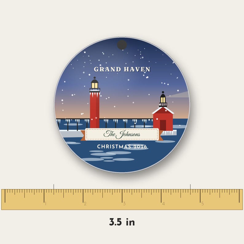 May include: A round, flat ornament featuring a winter scene of the Grand Haven lighthouse with red towers, a blue sky, and falling snow. A banner displays the text 'The Johnsons' and 'CHRISTMAS 2026'. A ruler shows a diameter of 3.5 inches.