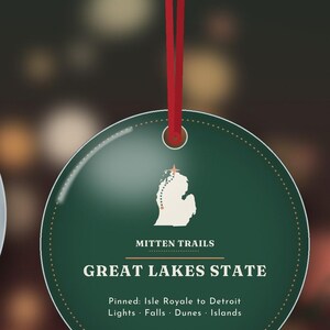 May include: A round dark green ornament hanging from a red ribbon. It features a white map of Michigan with a star, the text Mitten Trails, Great Lakes State, and Pinned: Isle Royale to Detroit, Lights, Falls, Dunes, Islands.