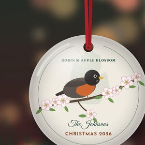 May include: A round white ceramic ornament hanging from a red ribbon. The design features a robin perched on a branch with pink apple blossoms. Text reads: ROBIN & APPLE BLOSSOM, The Johnsons, and CHRISTMAS 2026. A small button labeled Personalize is visible.