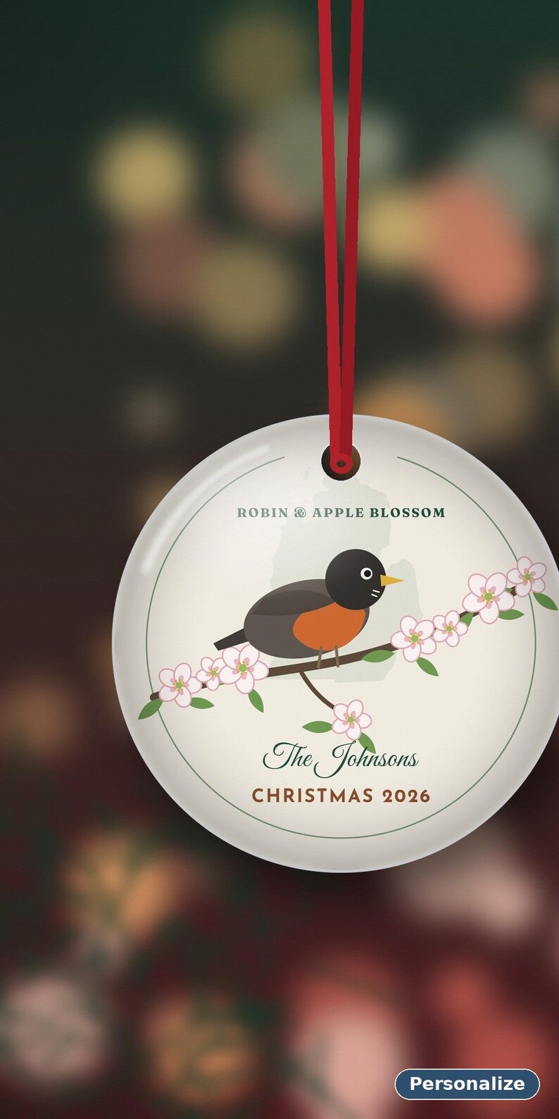 May include: A round white ceramic ornament hanging from a red ribbon. The design features a robin perched on a branch with pink apple blossoms. Text reads: ROBIN & APPLE BLOSSOM, The Johnsons, and CHRISTMAS 2026. A small button labeled Personalize is visible.