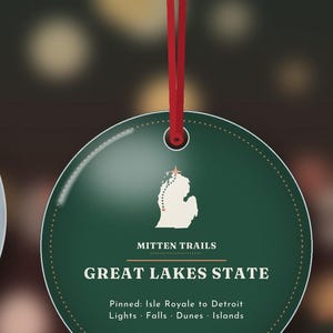 May include: A round, dark green ornament hanging from a red ribbon. The centre features a white map of Michigan with a star, the text MITTEN TRAILS, GREAT LAKES STATE, and Pinned: Isle Royale to Detroit Lights &middot; Falls &middot; Dunes &middot; Islands.