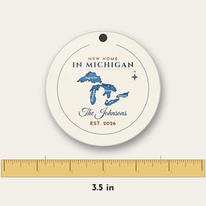 May include: A round, cream-colored ornament featuring a blue map of the Great Lakes, the text "NEW HOME IN MICHIGAN", "The Johnsons", and "EST. 2026". A ruler below shows the ornament has a diameter of 3.5 inches.