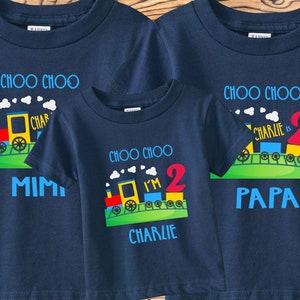 May include: Three navy blue t-shirts with a train design and the text "Choo Choo". The smallest shirt says "I'm 2 Charlie" and the middle shirt says "Charlie is 2". The largest shirt says "Papa".