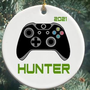 May include: White ceramic Christmas ornament with a black video game controller and the text "HUNTER" in green. The year "2021" is printed in green at the top of the ornament.