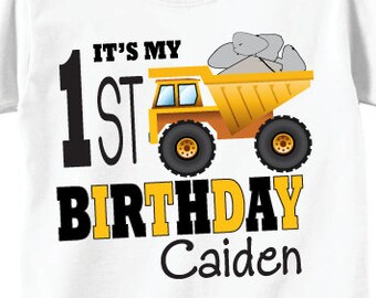 May include: White t-shirt with a yellow dump truck graphic carrying gray rocks, and the text "IT'S MY 1ST BIRTHDAY Caiden". The back of the shirt features a large number 1 with black and yellow stripes and a dump truck.