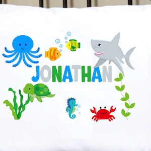 May include: White pillowcase featuring a colorful under-the-sea design. The design includes a blue octopus, a gray shark, a green turtle, and the name "JONATHAN" in colorful letters. Other sea creatures include a seahorse, fish, and a red crab.