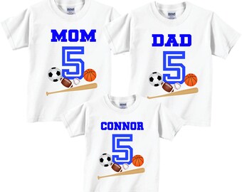 Op de afbeelding: Drie witte T-shirts met blauwe tekst en afbeeldingen. De shirts zeggen "Mam 5", "Pap 5" en "Connor 5". Elk shirt heeft een honkbalknuppel en een aantal sportballen, waaronder een voetbal, een rugbybal en een basketbal.
