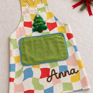 May include: A kitchen apron featuring a multicoloured wavy checkerboard pattern with yellow straps. It includes a green plush Christmas tree patch with the text Merry Christmas, a removable green towel pocket, and the name Anna in green and red rope.
