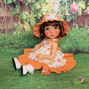 May include: A doll wearing a white dress with orange polka dots and a mushroom print. The doll is wearing a matching orange and white sun hat with a white ruffle. The doll is sitting on a green grassy surface.