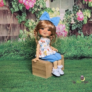 May include: A doll with blonde hair wearing a blue and white polka dot dress with a blue bow in her hair. She is sitting on a wooden box in front of a green grassy area.