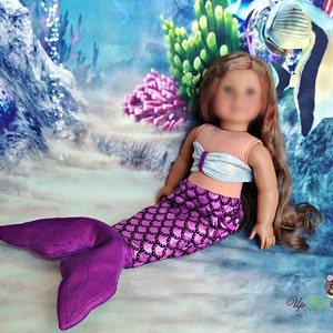 May include: A doll wearing a mermaid costume with a pink and purple tail and a silver and peach top. The doll is sitting on a background that looks like an underwater scene.