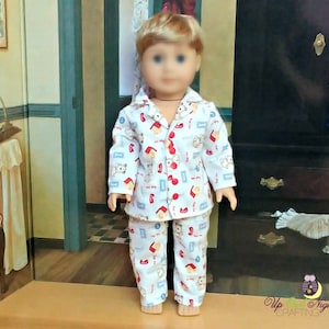 May include: A doll wearing a white pajama set with a repeating pattern of red houses, red hearts, and gray bears. The pajama top has red buttons and a collar. The pajama pants have an elastic waistband. The doll has blonde hair and blue eyes.