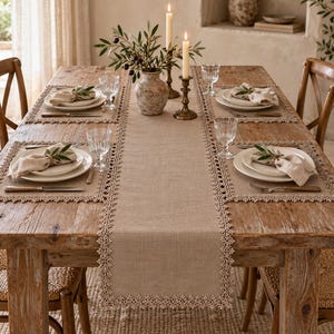 May include: A rectangular beige linen table runner with intricate lace trim edges, centered on a wooden dining table. The setting includes four place mats, white ceramic plates, linen napkins with olive sprigs, glassware, and two candles.