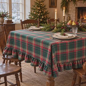 May include: A rectangular tablecloth featuring a green, red, and tan plaid pattern with a ruffled edge. The fabric covers a dining table set with white plates, folded napkins, silver cutlery, and two tall candles in brass holders.