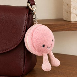 以下が含まれることがあります： 笑顔の顔、黒い刺繍の目、曲線的な口を持つピンク色のマカロンのぬいぐるみキーホルダー。柔らかく丸いマカロンにはクリーム色の中心層があり、銀色の金属製ロブスタークラスプに2本のぶら下がったリブ編みのピンク色の脚が付いています。