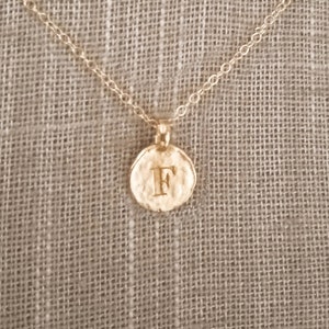 May include: A gold-colored necklace with a delicate chain and a circular pendant. The pendant features the letter "F" in a textured, embossed design. The necklace is displayed on a neutral-toned mannequin.