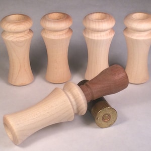 May include: A collection of wooden duck call parts. The calls are made of light-colored wood with a flared body and a rounded top. One call is assembled with a darker wood mouthpiece. Two brass shell casings are also present.