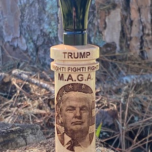 May include: A wooden duck call with a black top. The call is engraved with the text "TRUMP", "FIGHT! FIGHT! FIGHT!" and "M.A.G.A."  A portrait of Donald Trump is engraved on the call. The call also has the year "2024" engraved on it.