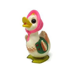 May include: A white duck figurine with a pink bonnet and green vest. The duck has a yellow beak and black eyes. The figurine is standing on a yellow base.