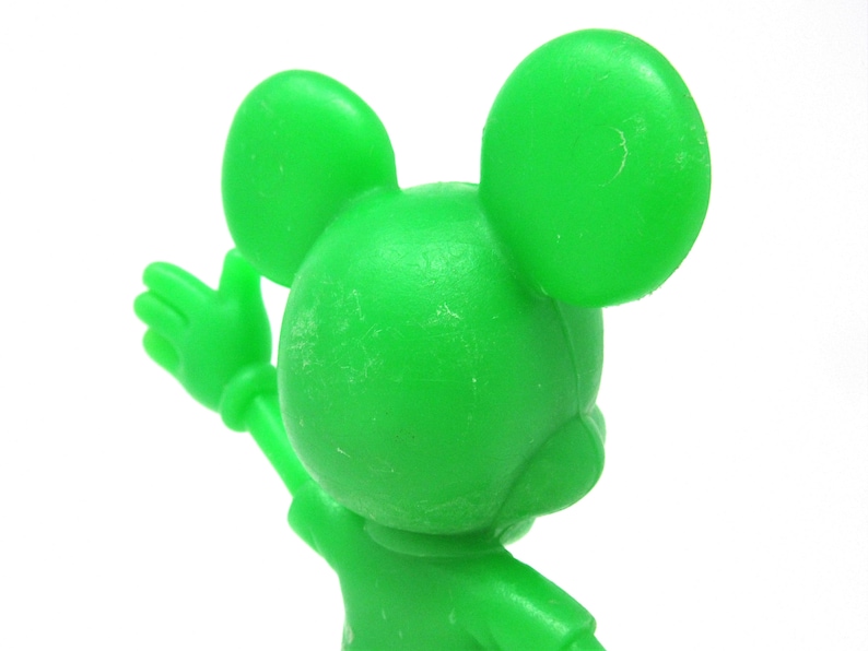 May include: A green plastic figurine of Mickey Mouse with his arms raised. The figurine is facing away from the camera.