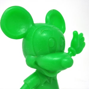 May include: A green plastic figurine of Mickey Mouse waving with his right hand. The figurine is a cartoon representation of the famous Disney character.