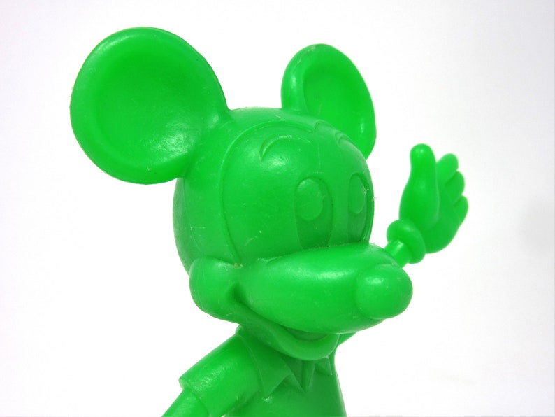 May include: A green plastic figurine of Mickey Mouse waving with his right hand. The figurine is a cartoon representation of the famous Disney character.