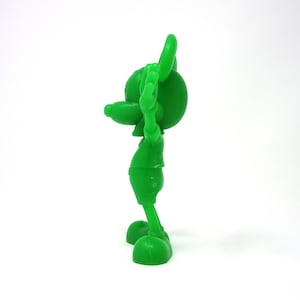 May include: A green plastic figurine of Mickey Mouse with his arms raised in the air. The figurine is standing on a white surface.