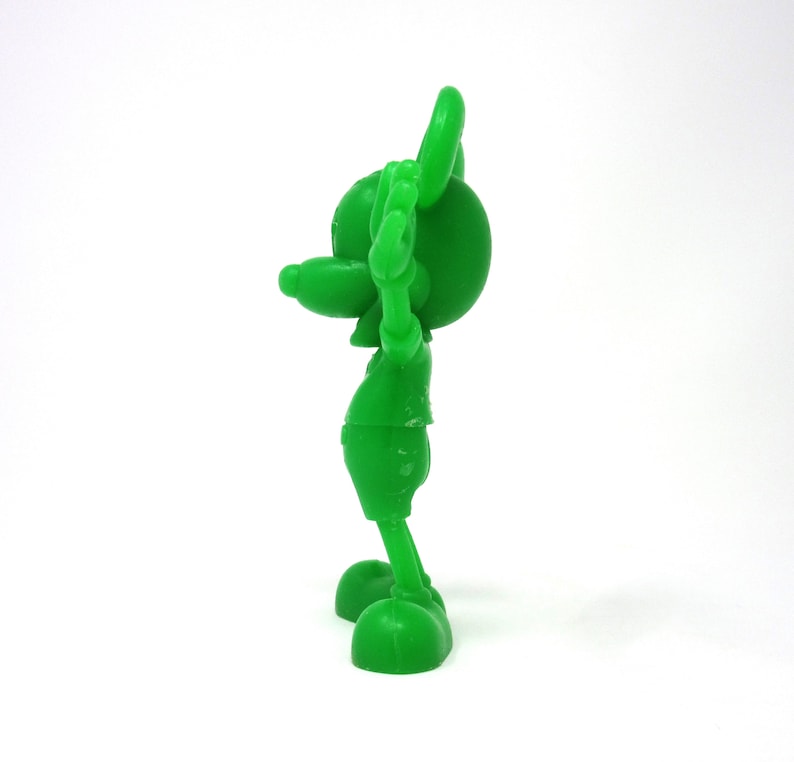 May include: A green plastic figurine of Mickey Mouse with his arms raised in the air. The figurine is standing on a white surface.