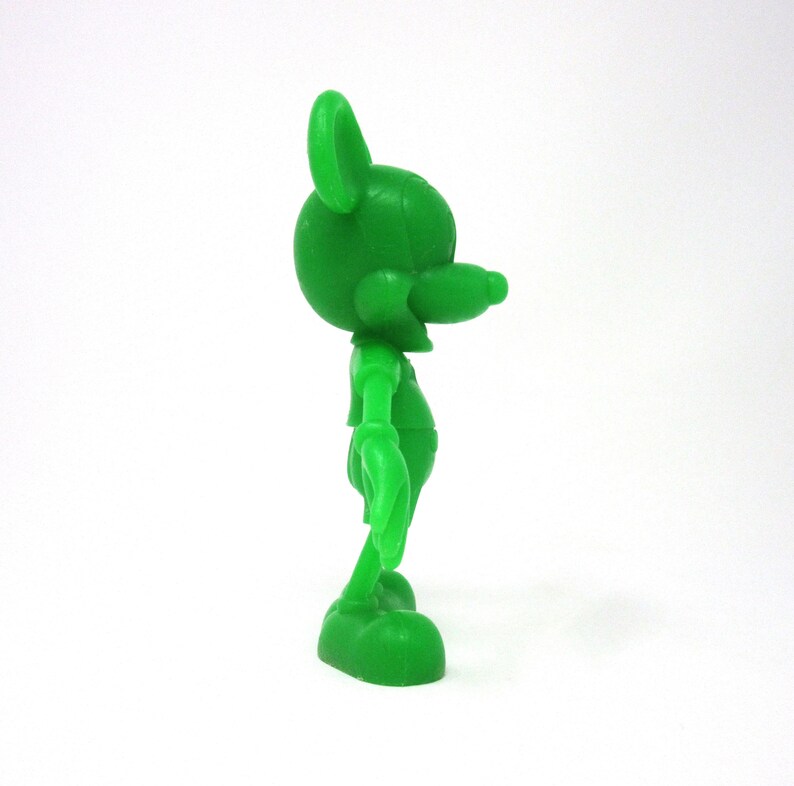 May include: A green plastic figurine of Mickey Mouse standing with his right arm raised and his left arm at his side. The figurine is facing to the right.