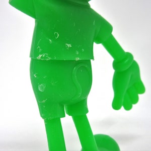 May include: A green plastic toy figure of a cartoon character. The figure is wearing a short-sleeved shirt and shorts. The figure has a large head and small body. The figure is missing some paint.