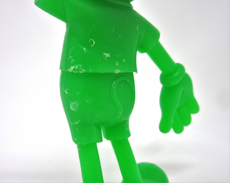 May include: A green plastic toy figure of a cartoon character. The figure is wearing a short-sleeved shirt and shorts. The figure has a large head and small body. The figure is missing some paint.