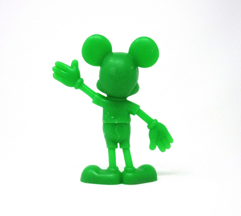 May include: A green plastic figurine of Mickey Mouse with his right arm raised in a wave. He is wearing shorts and has a friendly expression.