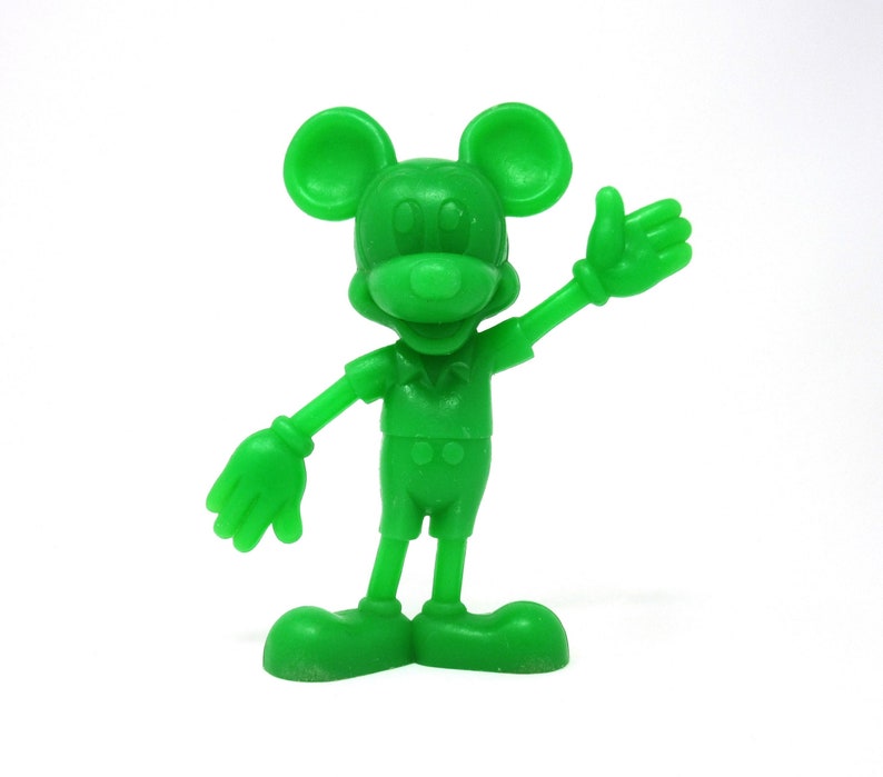 May include: A green plastic figurine of Mickey Mouse with a friendly expression. He is wearing a short-sleeved shirt and shorts. He is waving with his right hand.