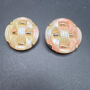 May include: A pair of round, clip-on earrings. The earrings have a woven design in shades of peach, cream, and gold. Gold-colored rectangular accents are placed around the edges of the earrings. The earrings are set against a dark gray background.