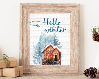 Puede incluir: Una pintura de acuarela enmarcada de una cabaña acogedora en la nieve con un abeto azul en el fondo. El texto "Hello winter" está escrito en cursiva azul sobre la cabaña.