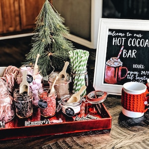 May include: A red tray with various ingredients for making hot cocoa, including marshmallows, chocolate chips, peppermint sprinkles, and sea salt. The tray is decorated with Hershey's chocolate bars and a small Christmas tree. A sign reads "Welcome to our HOT COCOA BAR" with the hours "open 24/7 til Santa visits".