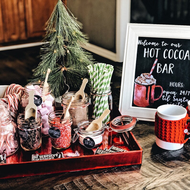 May include: A red tray with various ingredients for making hot cocoa, including marshmallows, chocolate chips, peppermint sprinkles, and sea salt. The tray is decorated with Hershey's chocolate bars and a small Christmas tree. A sign reads "Welcome to our HOT COCOA BAR" with the hours "open 24/7 til Santa visits".