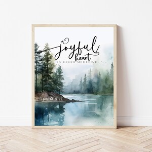 May include: A watercolor painting of a forest scene with a lake in the foreground. The text "A joyful heart is good medicine" is written in black cursive script. The text "Proverbs 17:22" is written in smaller black script below the main text.