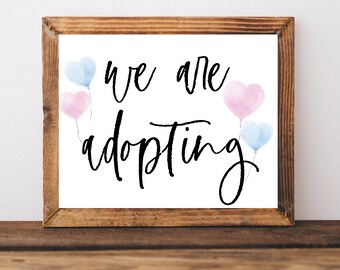 May include: A white sign with a wooden frame, featuring the text "We are adopting" in black cursive script. The sign is decorated with pink and blue watercolor heart-shaped balloons.