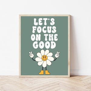 以下が含まれることがあります： 緑色のポスターに、レトロなスタイルの白い文字で「Let's Focus on the Good」と書かれています。テキストの下には、笑顔の顔とピースサインをした漫画のひまわりがあります。
