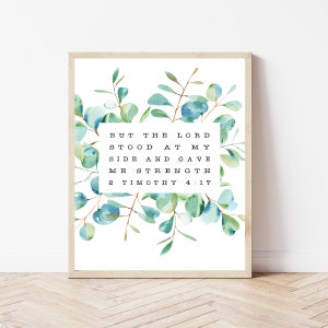 May include: A framed print with a watercolor illustration of eucalyptus leaves in shades of blue and green. The print features the Bible verse "But the Lord stood at my side and gave me strength. 2 Timothy 4:17".