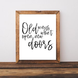 May include: A framed print with the quote "Old ways won't open new doors" in black script. The frame is made of light brown wood, and the print is on a white background. The print is displayed on a wooden surface.