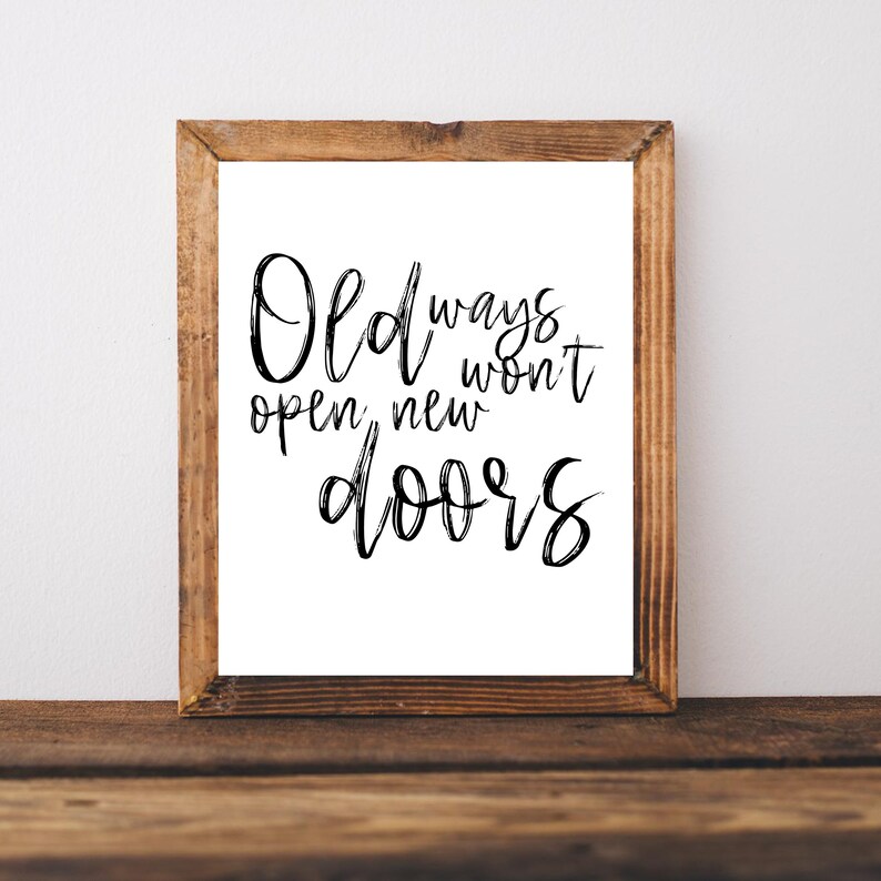 May include: A framed print with the quote "Old ways won't open new doors" in black script. The frame is made of light brown wood, and the print is on a white background. The print is displayed on a wooden surface.