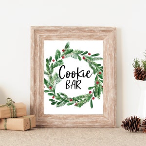 May include: A framed print with a rustic wooden frame. The print features the words "Cookie Bar" in black script, surrounded by a watercolor wreath of green pine needles, leaves, and red berries. The background is white.