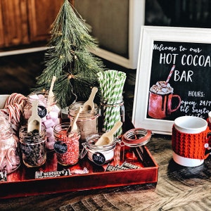 May include: A festive hot cocoa bar setup with various toppings in glass jars, including marshmallows, chocolate chips, and peppermint. A chalkboard sign reads "Hot Cocoa Bar." A mug with a red knitted cozy sits nearby. A small Christmas tree adds to the holiday theme.