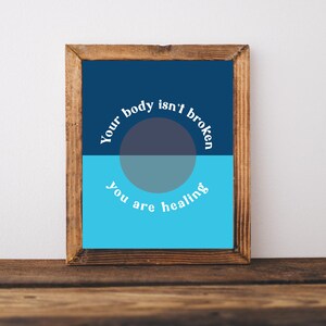 May include: A framed print with the text "Your body isn't broken, you are healing." The artwork features a two-tone design with a dark blue upper half and a light blue lower half, with a gray circle in the center.
