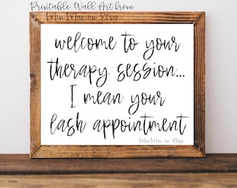 May include: A white framed print with black text that reads "Welcome to your therapy session... I mean your lash appointment." The text is in a handwritten style font. The print is on a wooden surface.