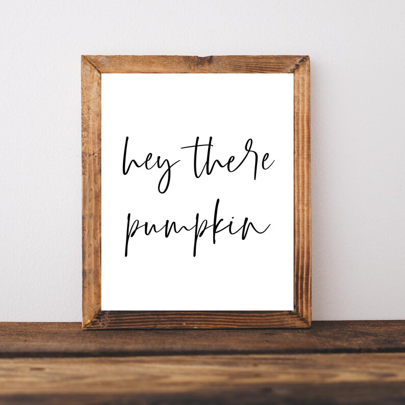 May include: A framed print with the words "hey there pumpkin" in black script. The print is set in a rustic, wooden frame, and rests on a wooden surface. The background is a plain white wall.